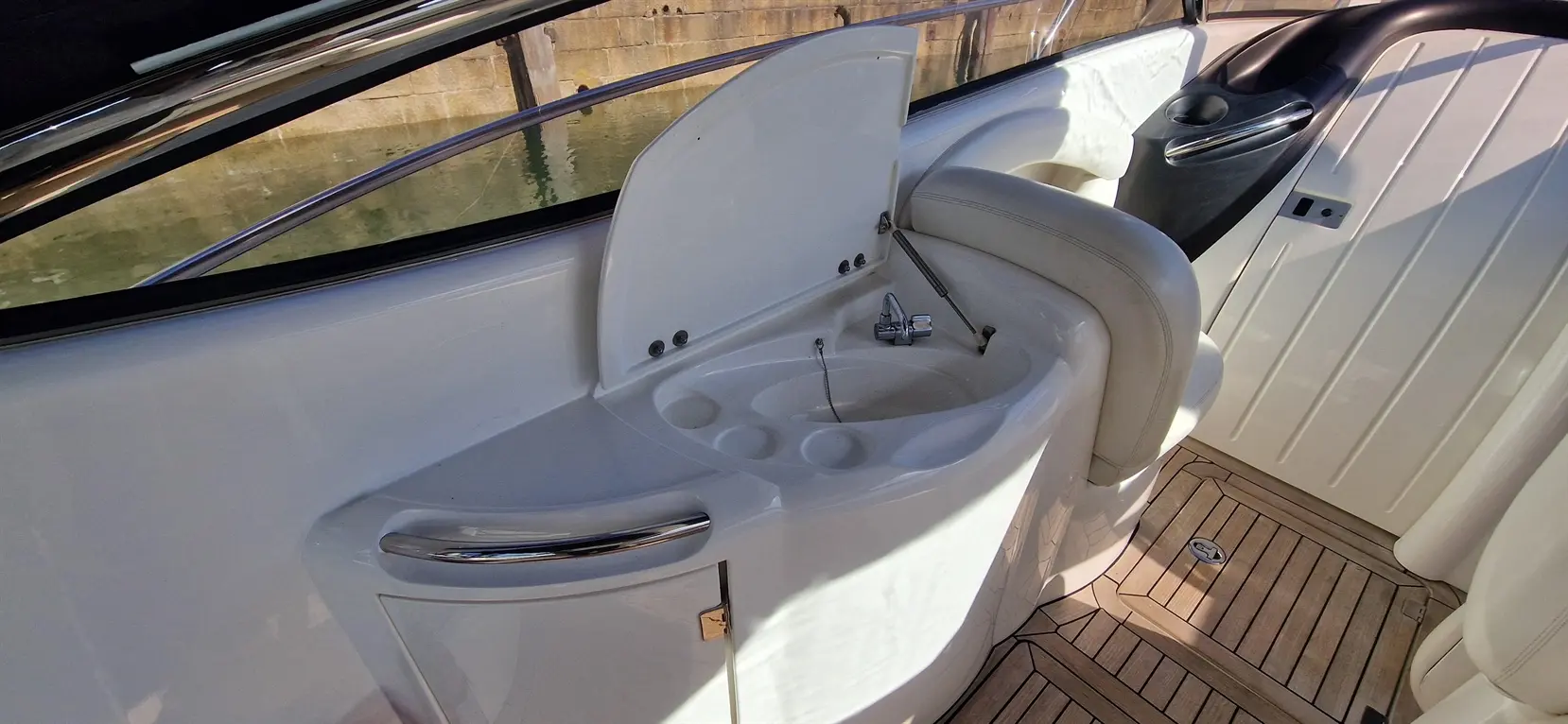 Interior of a boat featuring a sink, with a chrome faucet, next to cushioned seating and wooden flooring.